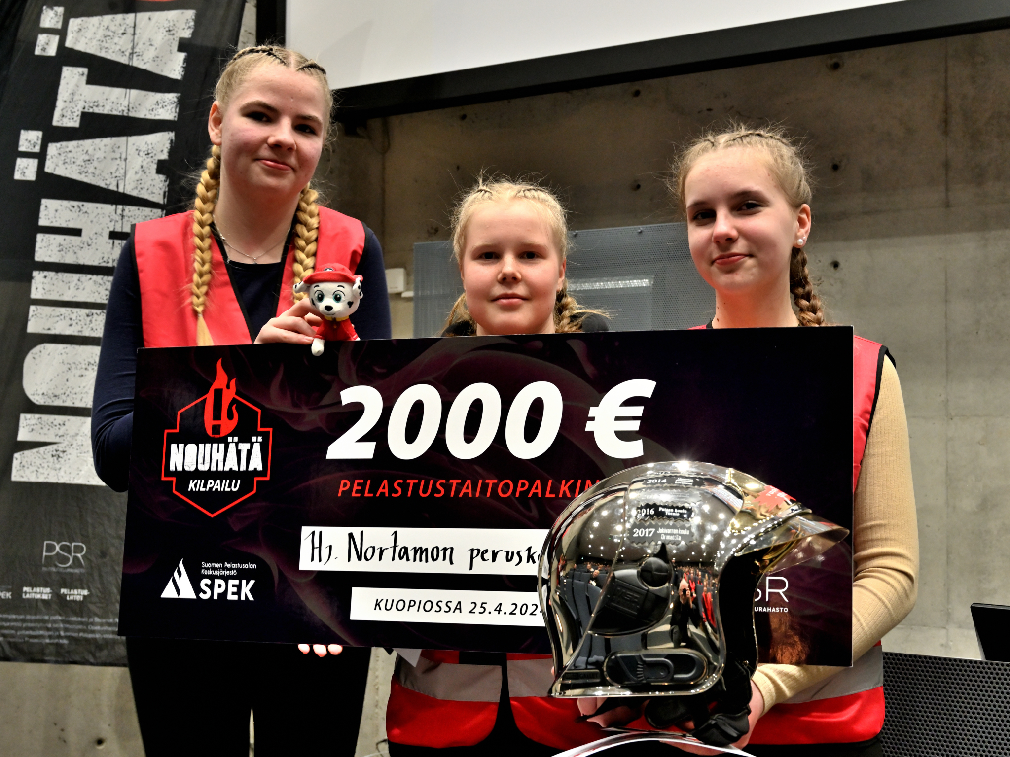 Kolme lettipäistä tyttöä pitävät kädessään kilpailut voittosekkiä ja kultaista palomiehen kypäerää.